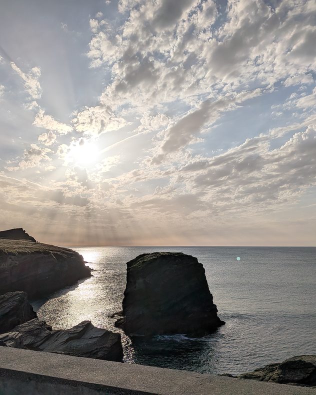Bright sun shining through scattered altocumulus clouds above a rocky coastline, with crepuscular rays illuminating the calm sea, rugged cliffs, and a large offshore sea stack.