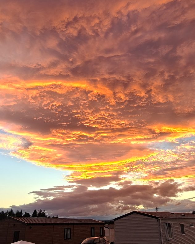 Dramatic orange and gold mammatus clouds glowing at sunset above rooftops, with pouch-shaped cloud formations, layered storm clouds, and vivid evening light illuminating the underside of the sky.